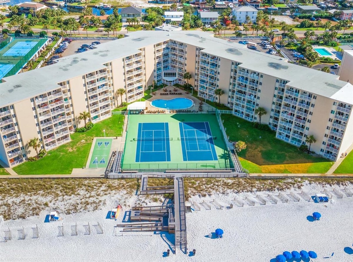 An aerial view of the condo complex displays multiple buildings surrounding well-maintained tennis courts and a central pool area. The adjacent beach features umbrellas and chairs, while lush greenery and pathways enhance the outdoor setting.