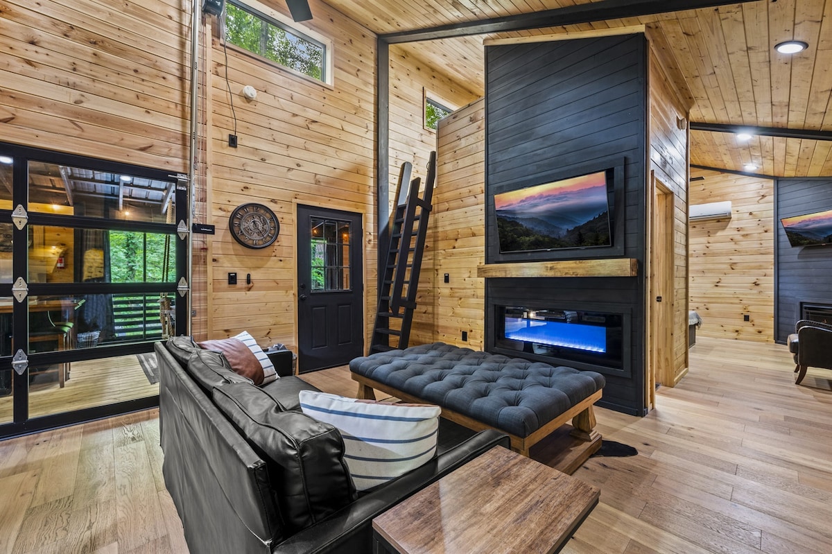 A spacious living area features warm wooden walls and modern furniture. A comfortable sofa sits adjacent to a large coffee table, while a sleek fireplace and a wall-mounted television add to the ambiance. A loft ladder can be seen, leading to additional sleeping space.