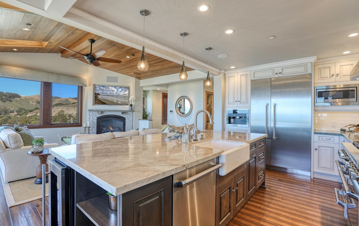 A spacious kitchen features a large island with a marble countertop, stainless steel appliances, and elegant cabinetry. Natural light streams through windows that offer views of the surrounding landscape, while wood accents in the ceiling add warmth to the modern design.