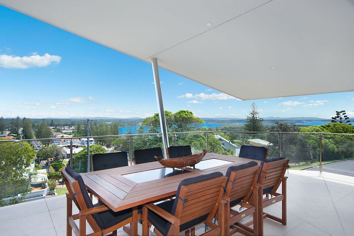 An outdoor dining area features a large wooden table surrounded by eight chairs, set against a backdrop of expansive blue skies and distant water views. Glass railings enhance visibility, allowing the serene landscape to be part of the experience.