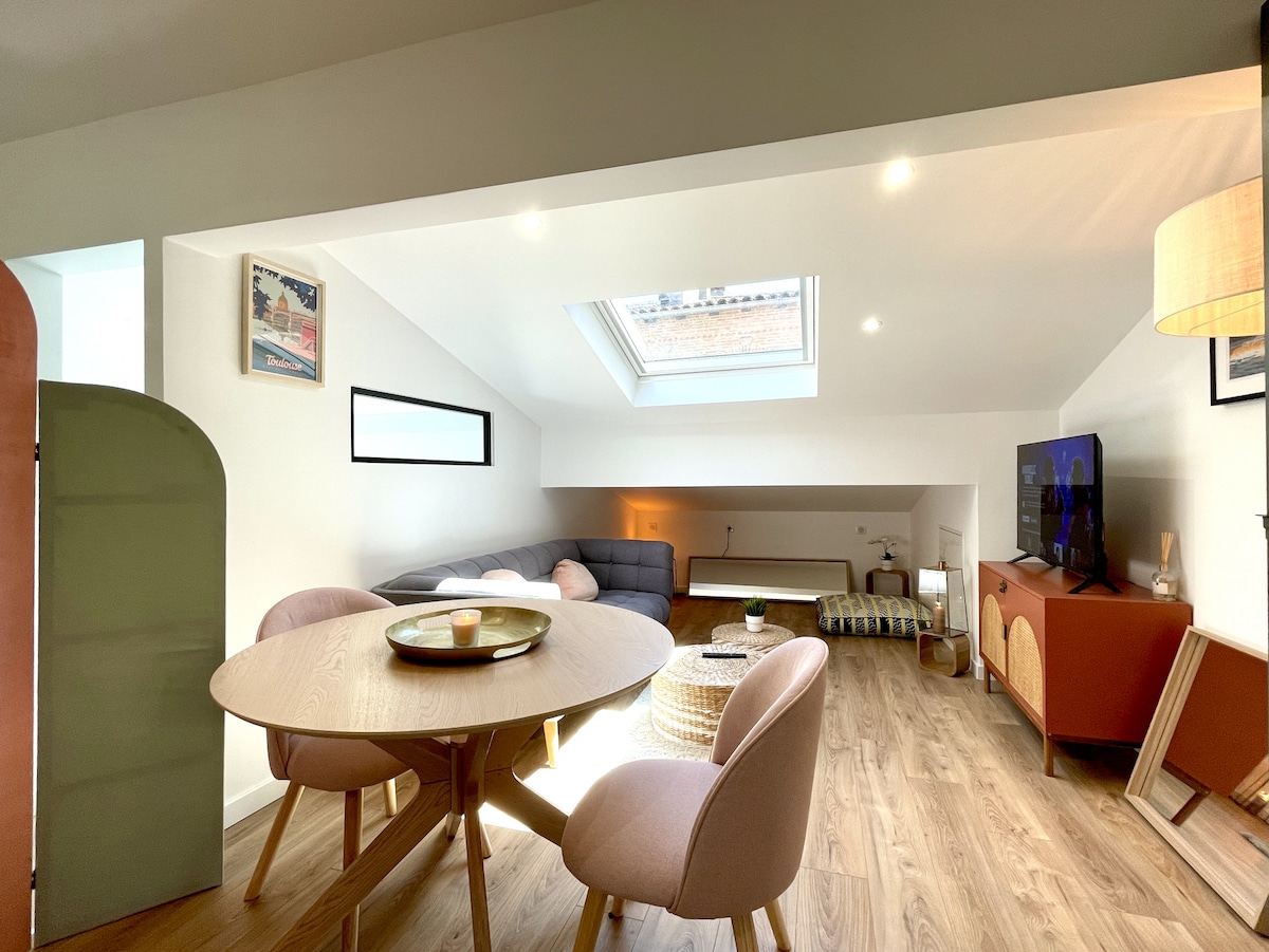 A bright living area features a cozy seating arrangement with a gray sofa and a low table. A round dining table for two is positioned nearby. Natural light floods the space through a skylight, illuminating the wooden floor and modern furnishings.