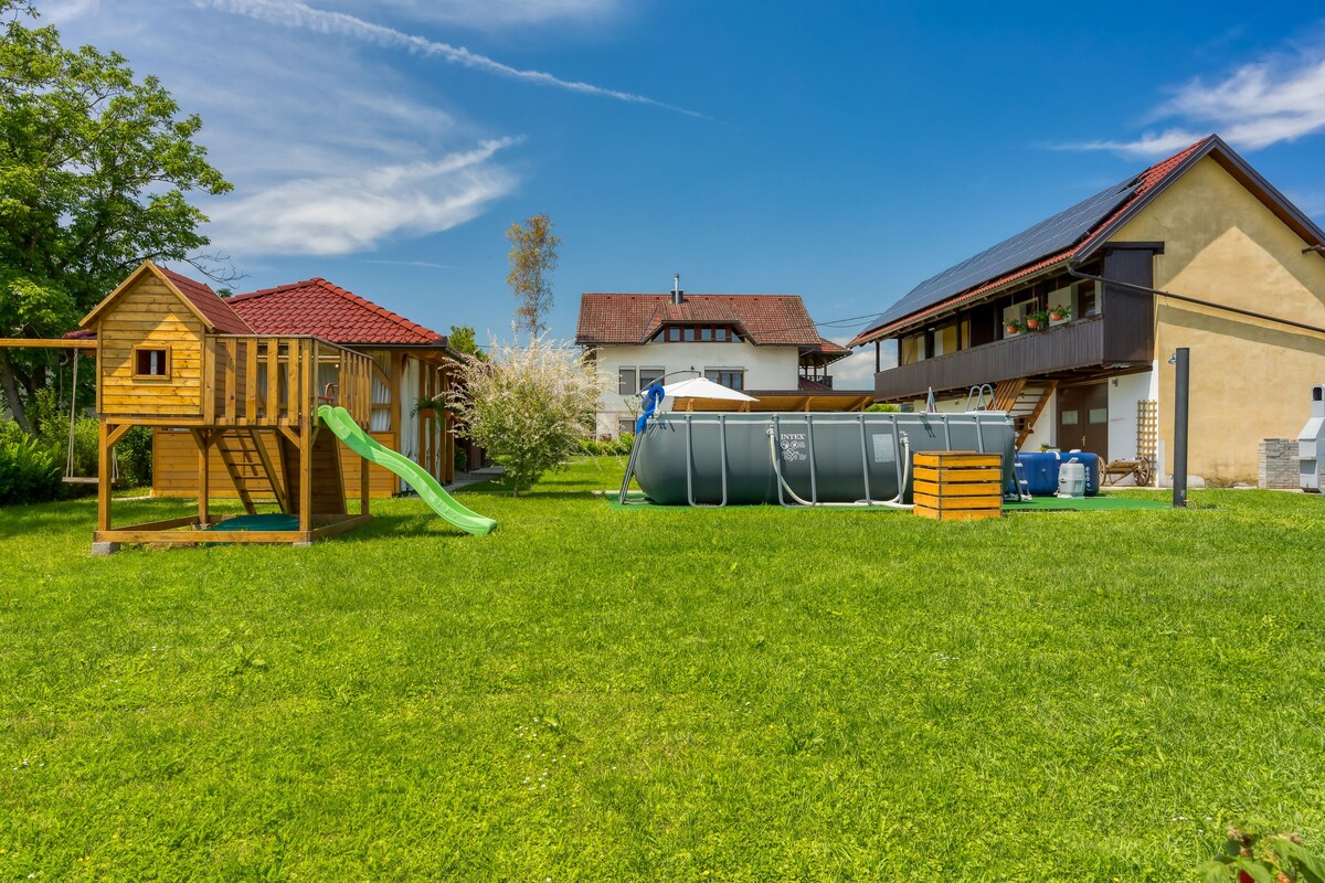 A well-maintained outdoor area features a wooden playhouse with a slide and a spacious grassy lawn. Adjacent to it, a circular above-ground pool is situated, along with an outdoor shower. The backdrop includes a large two-story house with multiple windows and a sloped roof.
