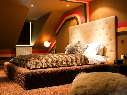 A uniquely styled bedroom is highlighted by bold colour stripes on the walls and a plush, textured floor. A spacious, upholstered bed is accompanied by decorative pillows and a cozy blanket, while ambient lighting creates a warm, relaxing environment.