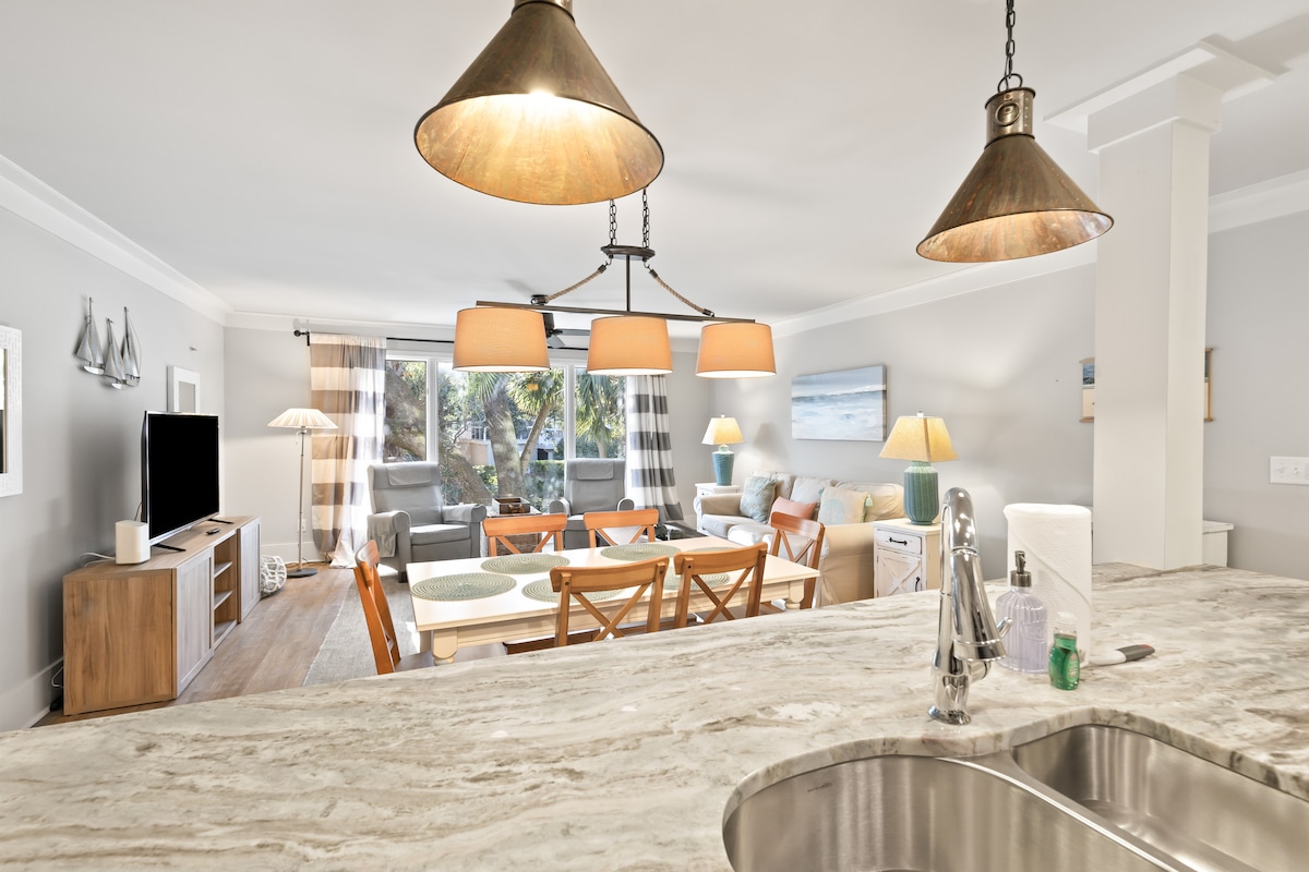 A spacious living area features a dining table set for six and comfortable seating arrangements. Soft lighting is provided by multiple pendant lights. The view through large windows reveals a bright outdoor space with lush greenery, enhancing the openness of the room.