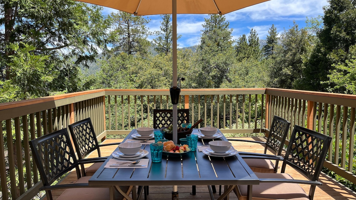 An outdoor dining area is set with a table and comfortable seating. Plates and turquoise glasses rest on the table, complemented by a platter of pastries. An umbrella provides shade, while the surrounding trees and blue sky enhance the natural ambiance.