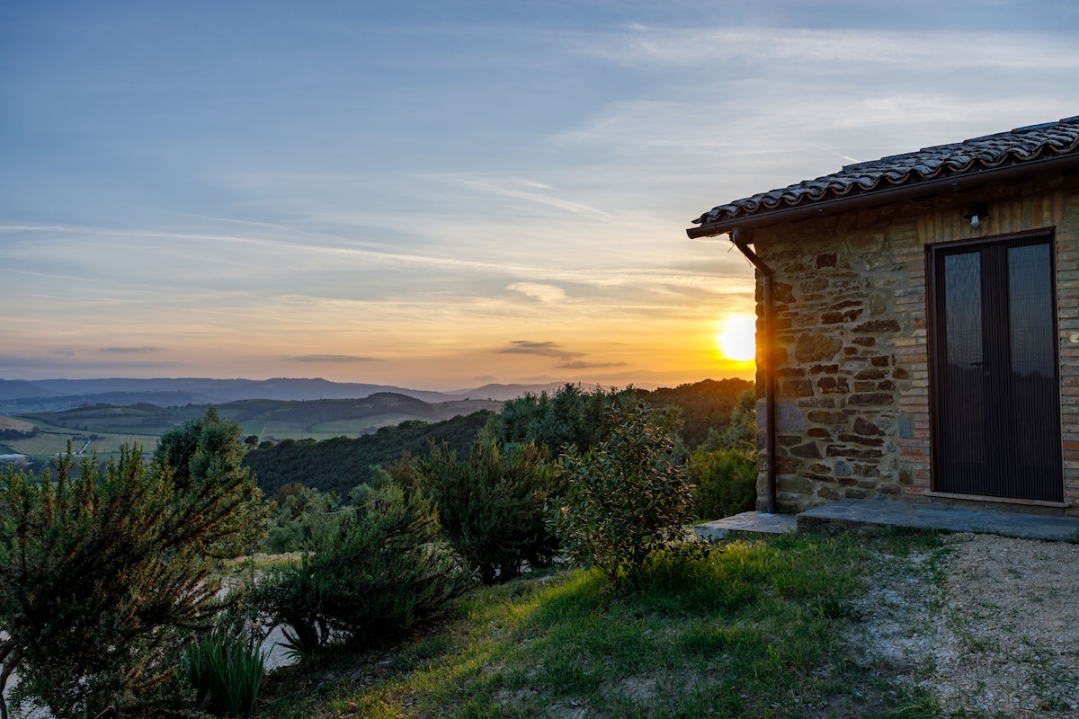 A rustic building is framed by lush greenery and gentle hills, with a golden sunset casting warm hues across the landscape. The natural stone and wooden elements harmonize with the surroundings, creating a serene outdoor environment.