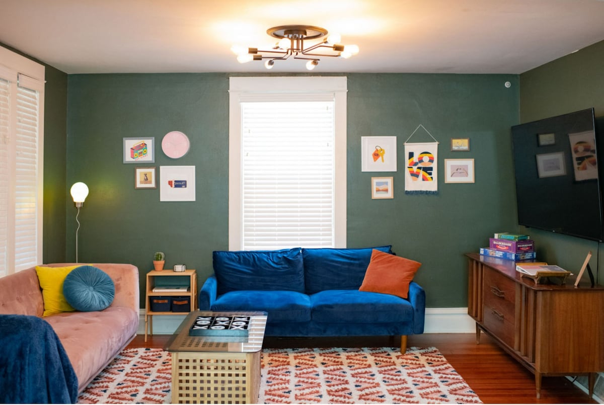 An eclectic living room is depicted with a pink couch and a blue couch, accented by colorful throw pillows. A patterned area rug covers the hardwood floor, while art is displayed on the green walls. A wooden media unit holds a TV, and a coffee table is center stage.
