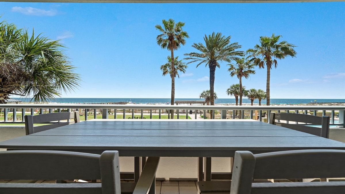 A spacious balcony table is set for leisure, overlooking palm trees and the serene beach. The ocean is visible in the distance, providing a vibrant backdrop to outdoor gatherings.