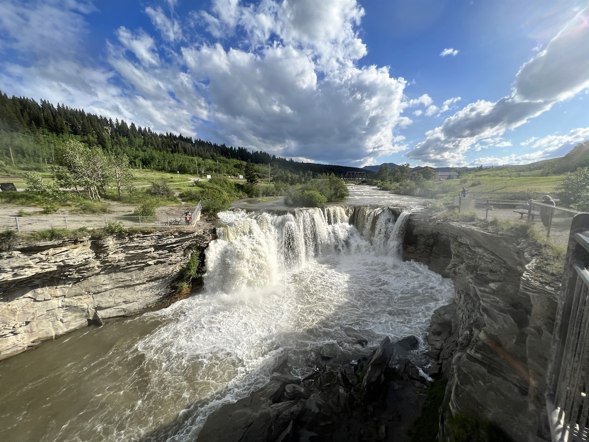 A waterfall cascades over layered rock formations into a flowing river below. Lush greenery surrounds the scene, with large clouds scattered above in a blue sky. The area appears accessible with walking paths visible alongside the water.