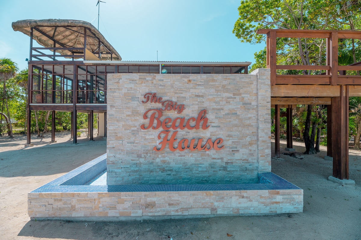 A modern beach house exterior is showcased, featuring a stone wall with the name 'The Big Beach House' prominently displayed. Large windows highlight the wooden framework above, surrounded by tropical vegetation and sandy pathways.