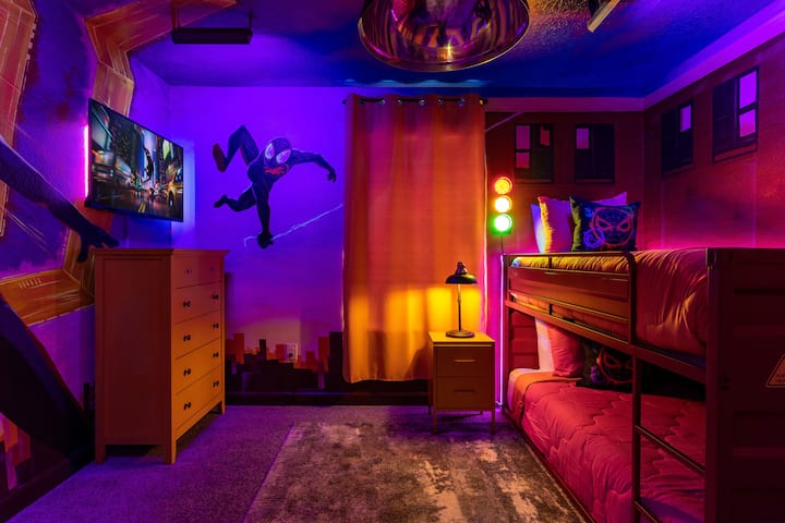 This room is a Spiderverse fan's dream come true, featuring twin-sized bunk beds, a wardrobe closet, and a TV. Completing the look are the vibrant city colors and the Spiderman mural on the wall.