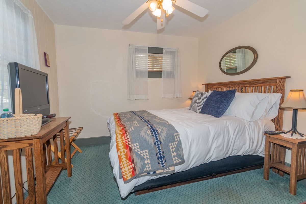 The bedroom features a spacious bed with layered white bedding and decorative pillows. A wooden headboard complements the warm tones of the room. A small television and a wicker basket are situated beside a wooden side table. Natural light filters in through sheer curtains, creating a calm ambiance.
