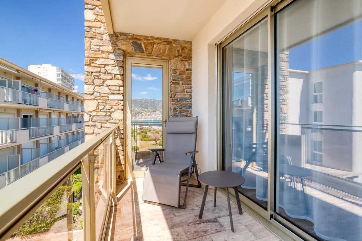 A sunny balcony features a comfortable lounge chair and a small table, surrounded by glass sliding doors. A view of the landscape and distant hills can be seen beyond the balcony railing, which is complemented by stone walls that add a natural texture to the space.