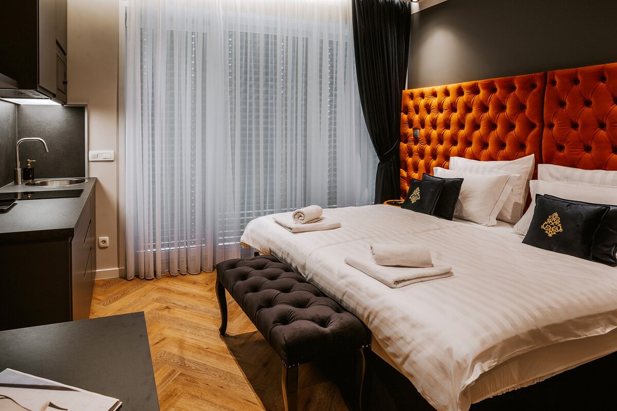 A modern bedroom features a luxuriously upholstered double bed with crisp linens and fluffy towels neatly arranged. A stylish bench sits at the foot of the bed. Natural light filters through sheer curtains, enhancing the warm wood flooring and chic decor.
