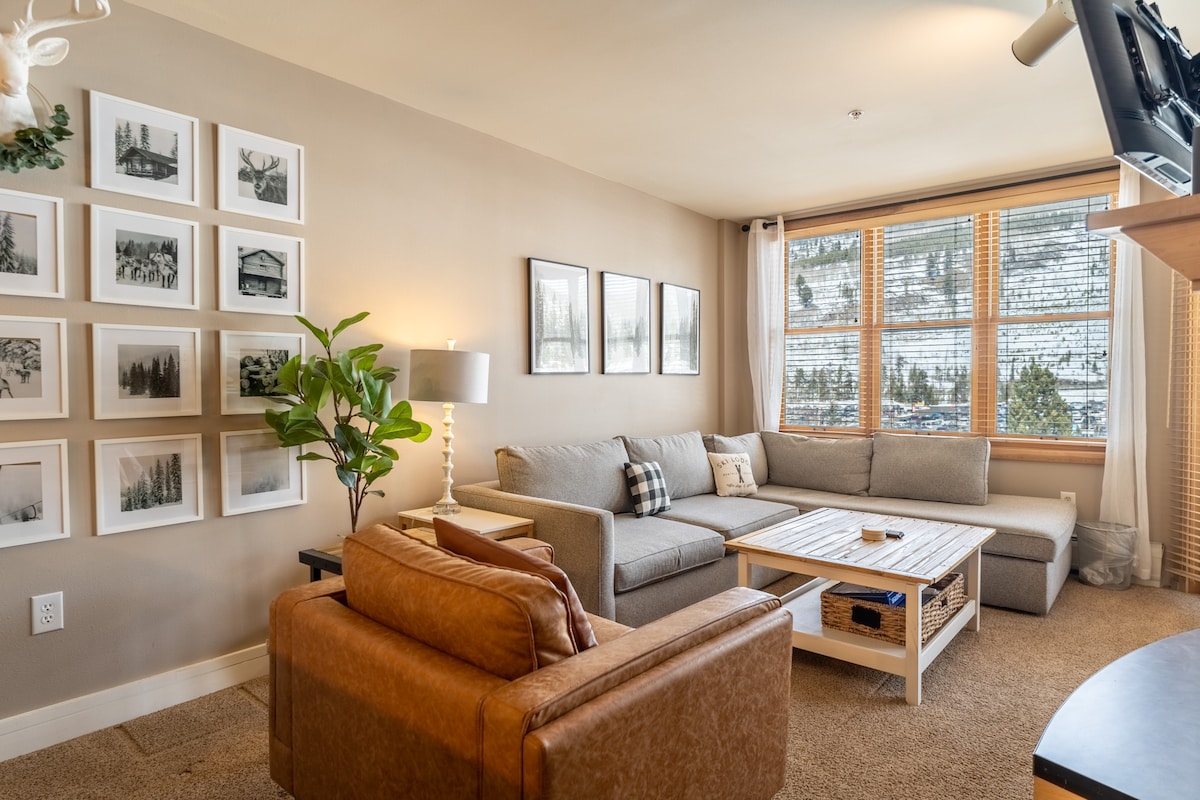 The living area features a large sectional sofa and a leather lounge chair, providing ample seating. A coffee table sits in the center, surrounded by framed artwork on the walls. Natural light streams in through the large window, showcasing views of snow-covered mountains.