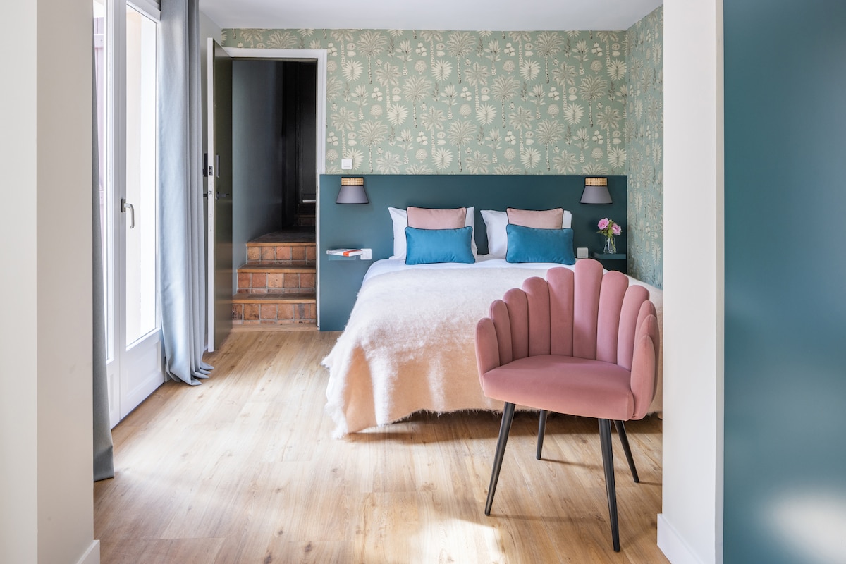 The suite features a spacious bed adorned with soft linens against a backdrop of nature-inspired wallpaper. A pink upholstered chair adds a touch of color, while natural light enters through large doors leading to a private terrace. Subtle wood tones enhance the floor design.