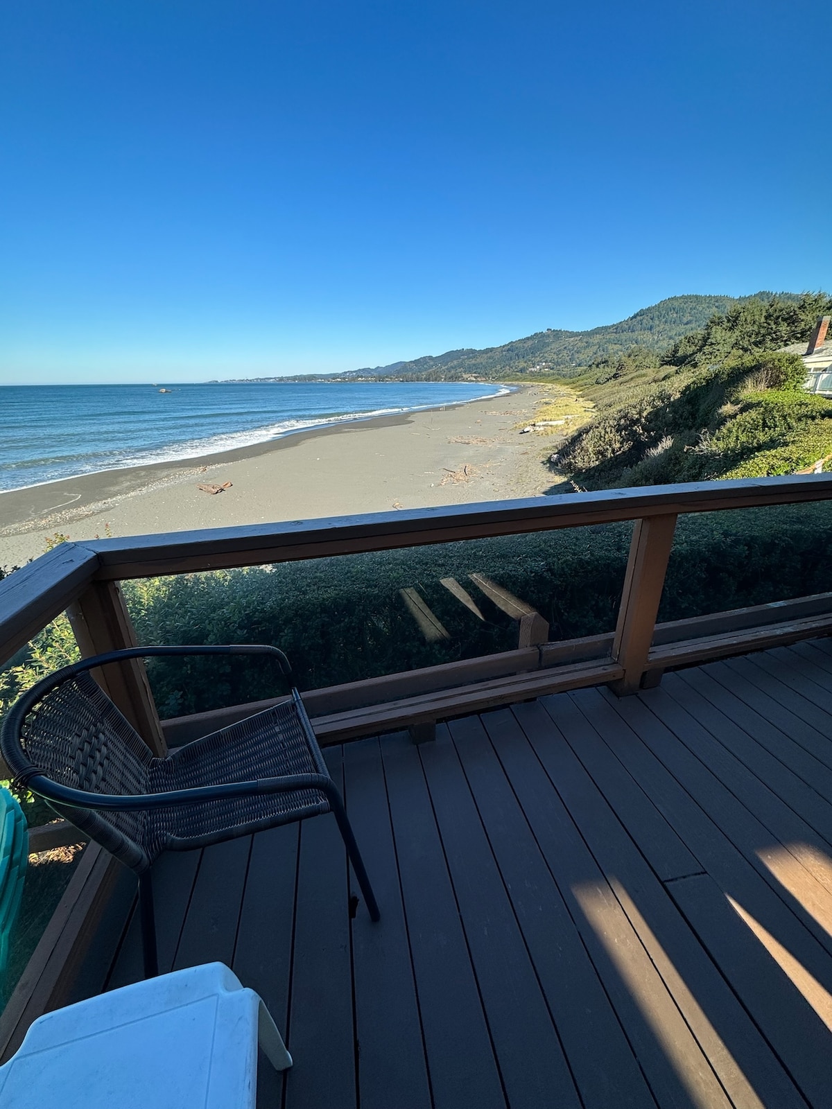The view from the deck overlooks a sandy beach and the ocean, surrounded by lush greenery and distant mountains under a clear blue sky. A weathered wood railing frames the scene, while a black wicker chair is positioned on the deck.