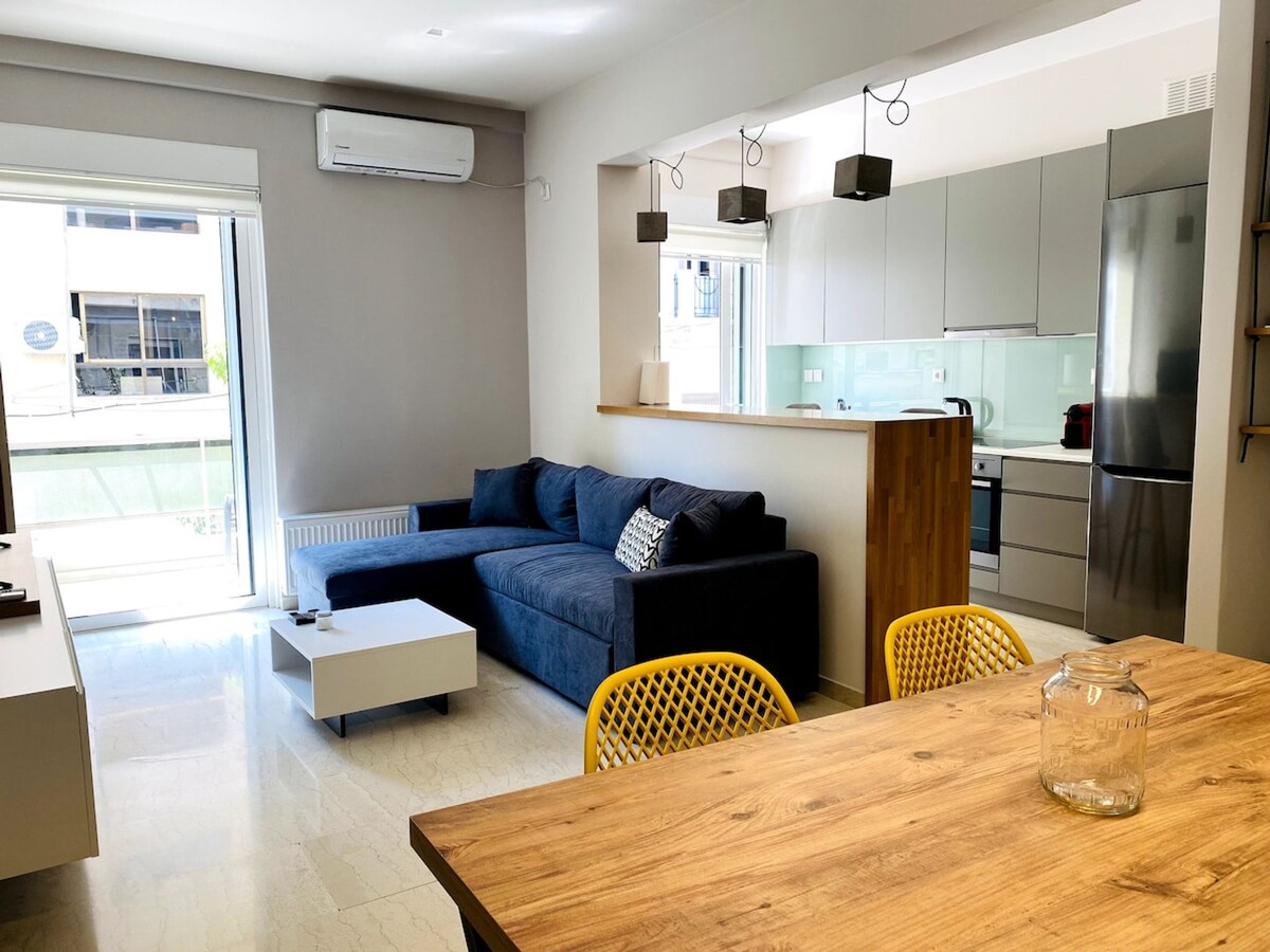 A modern living space features a spacious blue sofa accompanied by a light wood coffee table. A dining area with yellow chairs is situated next to a well-equipped kitchenette adorned with sleek cabinetry. Natural light filters in through large glass doors leading to a balcony.