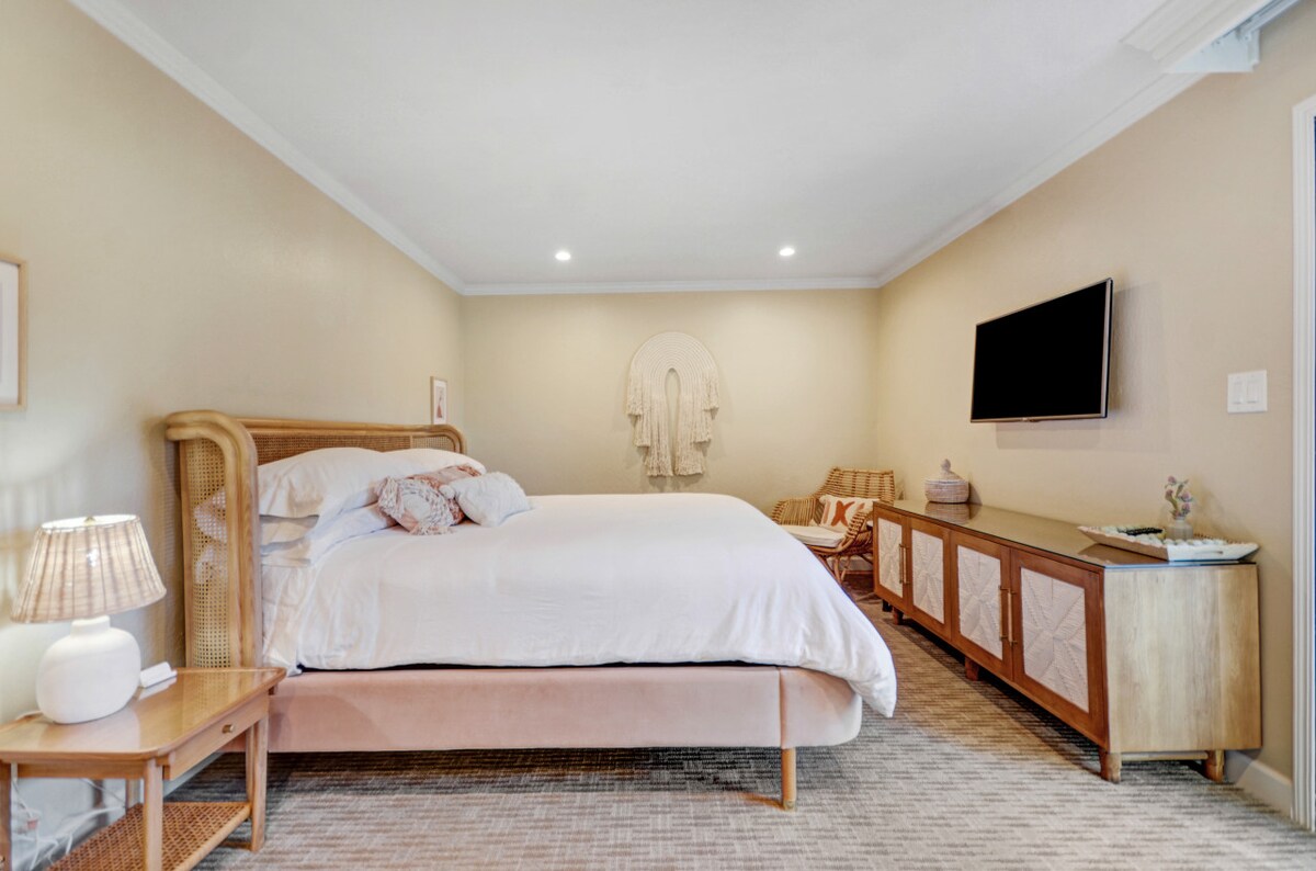 The bedroom features a cozy king bed dressed in soft linens. A comfortable chair is positioned next to a lighted bedside table, while a stylish dresser and a wall-mounted smart TV are present. Warm lighting creates a calming space.