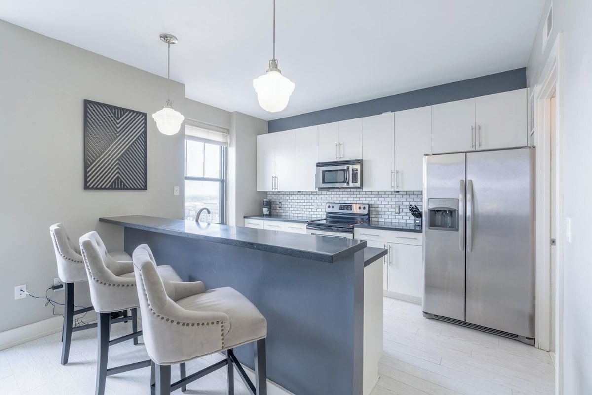A modern kitchen area features sleek countertops and stainless-steel appliances. Two bar stools are positioned at the kitchen island, providing space for meals or work. Large windows allow natural light to fill the room, highlighting the stylish cabinetry and backsplash.