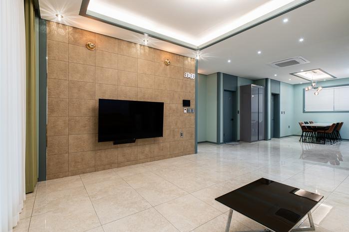 A spacious living area is highlighted by a large flat-screen television mounted on a textured wall. Neutral tile flooring reflects light from the ceiling fixtures, while an elegant dining area is visible in the background, featuring a rectangular table and several chairs.