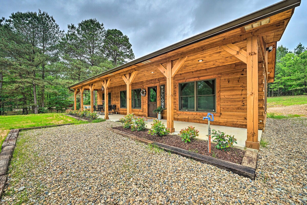 The rustic cabin showcases a log exterior, highlighting a spacious covered porch with wooden supports. Lush greenery surrounds the property, while a gravel pathway leads to the entrance. Flower beds add a touch of color, enhancing the serene natural setting.