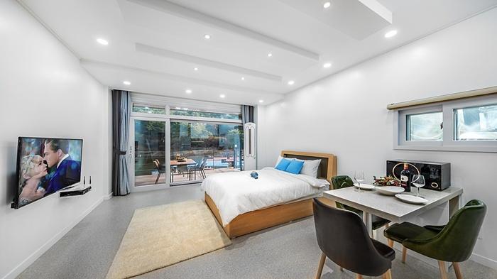 A spacious room features a large bed with decorative blue pillows and a cozy area rug. A dining table with chairs is positioned near a window, which overlooks a terrace area. Natural light illuminates the room, while a television provides entertainment.
