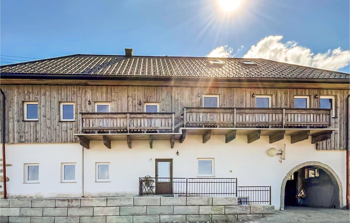 The exterior of the flat features a traditional wooden facade with balconies extending from the upper level. Sunlight shines brightly above, illuminating the inviting entrance and stone pathway leading to the door. A combination of rustic charm and modern design is evident.