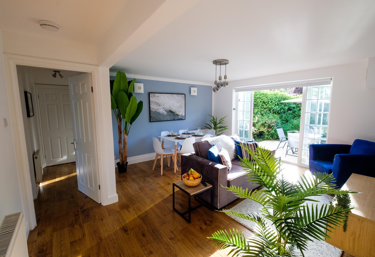 A bright living area showcases a mix of natural wood flooring and light-colored walls. A comfortable sofa sits opposite a coffee table, beside a plant. A dining area with multiple chairs is visible, and large glass doors open to a tranquil outdoor space.