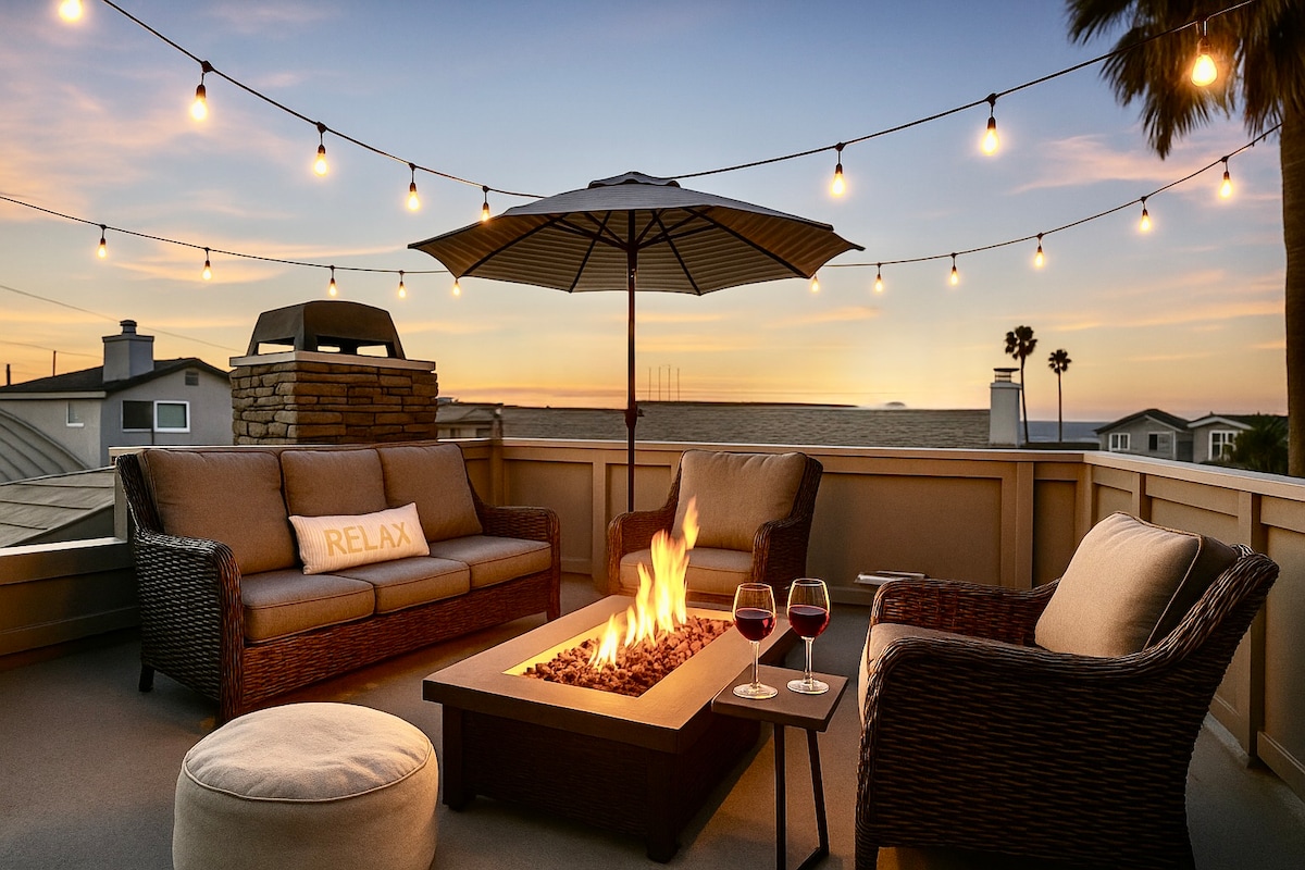 A rooftop deck is illuminated by string lights at sunset, featuring a cozy seating area with a fire pit in the center. Comfortable chairs are complemented by an umbrella, while a decorative cushion with the word 'RELAX' is positioned on a nearby seat.