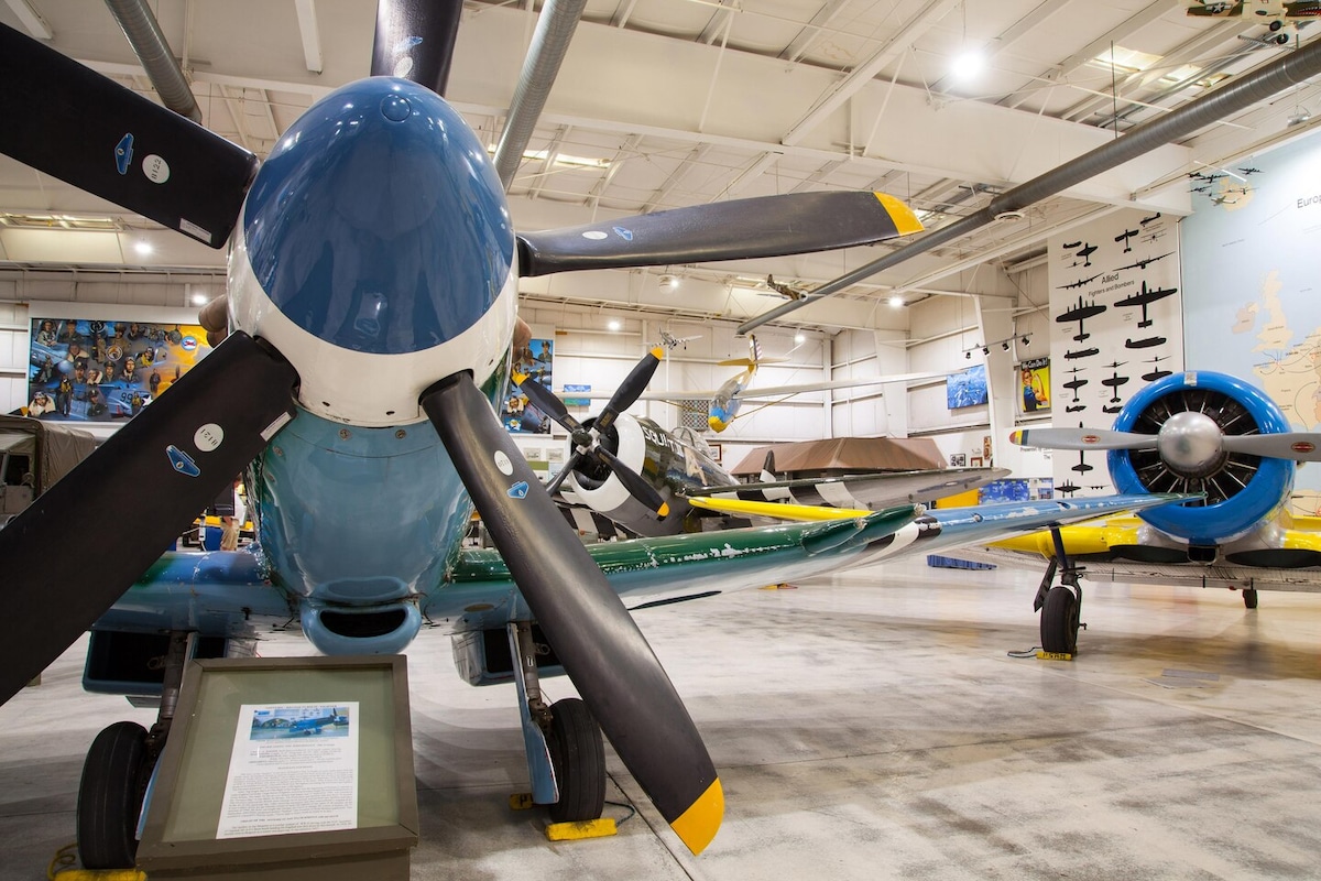 An exhibit featuring vintage aircraft is displayed in a spacious hangar. Prominent propeller blades in blue and black colors dominate the foreground. Various aircraft can be seen parked behind, with informative panels placed throughout the area.