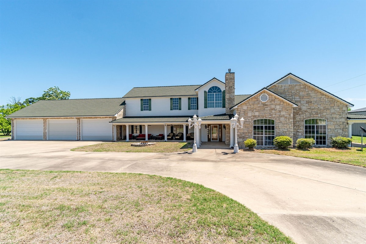 A spacious two-story home presents a mix of stone and siding exterior. A large driveway leads up to the entrance, framed by manicured lawns and flowering shrubs. Three vehicle bays are visible, along with a decorative entryway supported by white columns.