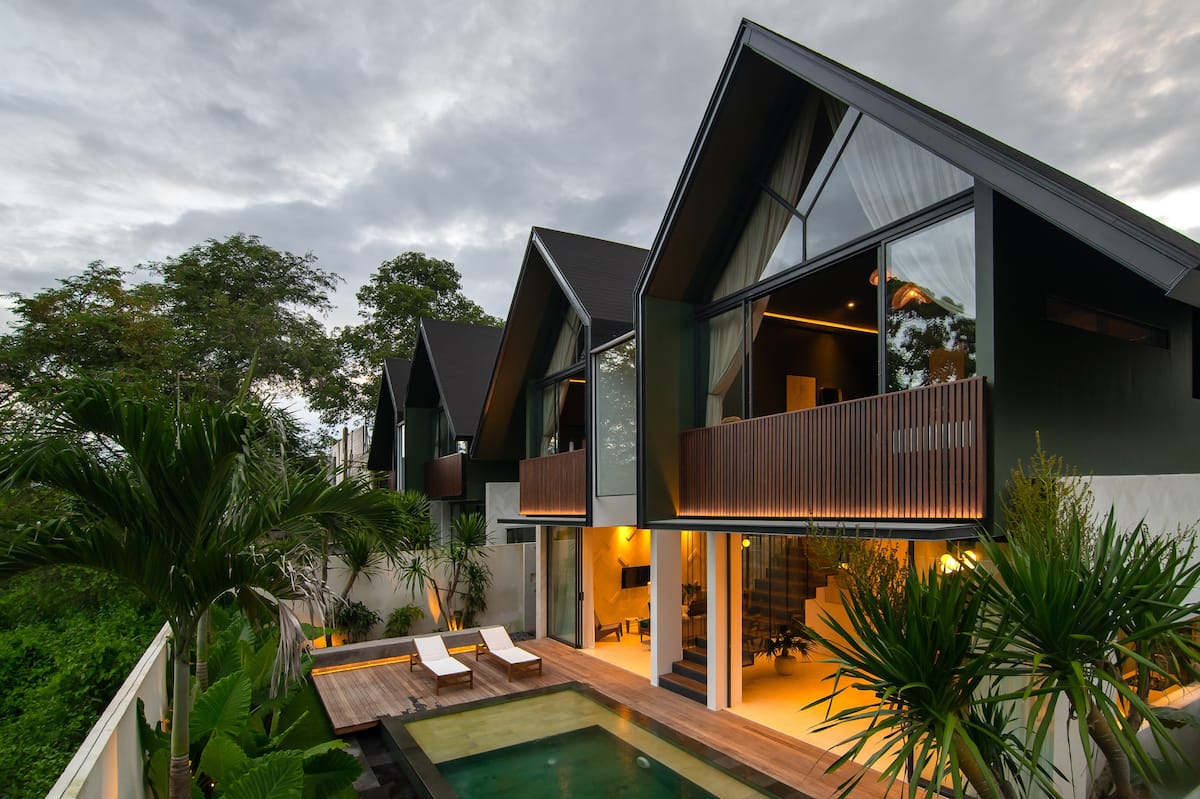 The exterior of the villa showcases a modern architectural design with multiple gabled rooftops. Large glass windows allow for natural light to enter the interior spaces, and a private pool is surrounded by a wooden deck and tropical greenery.