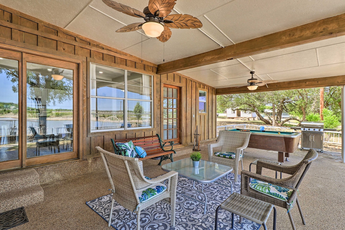 A covered patio is furnished with seating arrangements, including lounge chairs and a coffee table. Decorative pillows complement the space, while a hot tub is visible nearby. Large windows provide views of an outdoor area, enhancing the inviting atmosphere.