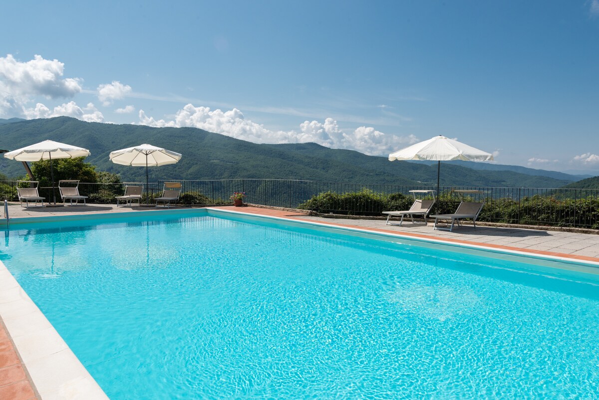A large swimming pool reflects the sky, surrounded by lounge chairs and umbrellas for shade. The pool area offers sweeping views of the mountainous landscape, with greenery surrounding the property. Soft clouds are visible above, creating a serene atmosphere.