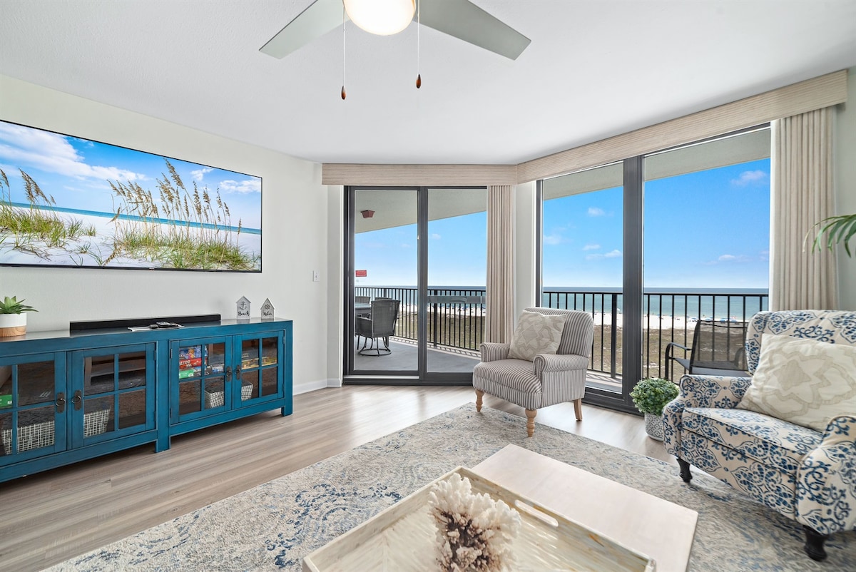 The living area features a large smart TV mounted on a white wall, complemented by a blue cabinet. Sliding glass doors offer access to a private balcony with a stunning ocean view. A cozy patterned armchair and a light-colored area rug complete the inviting space.