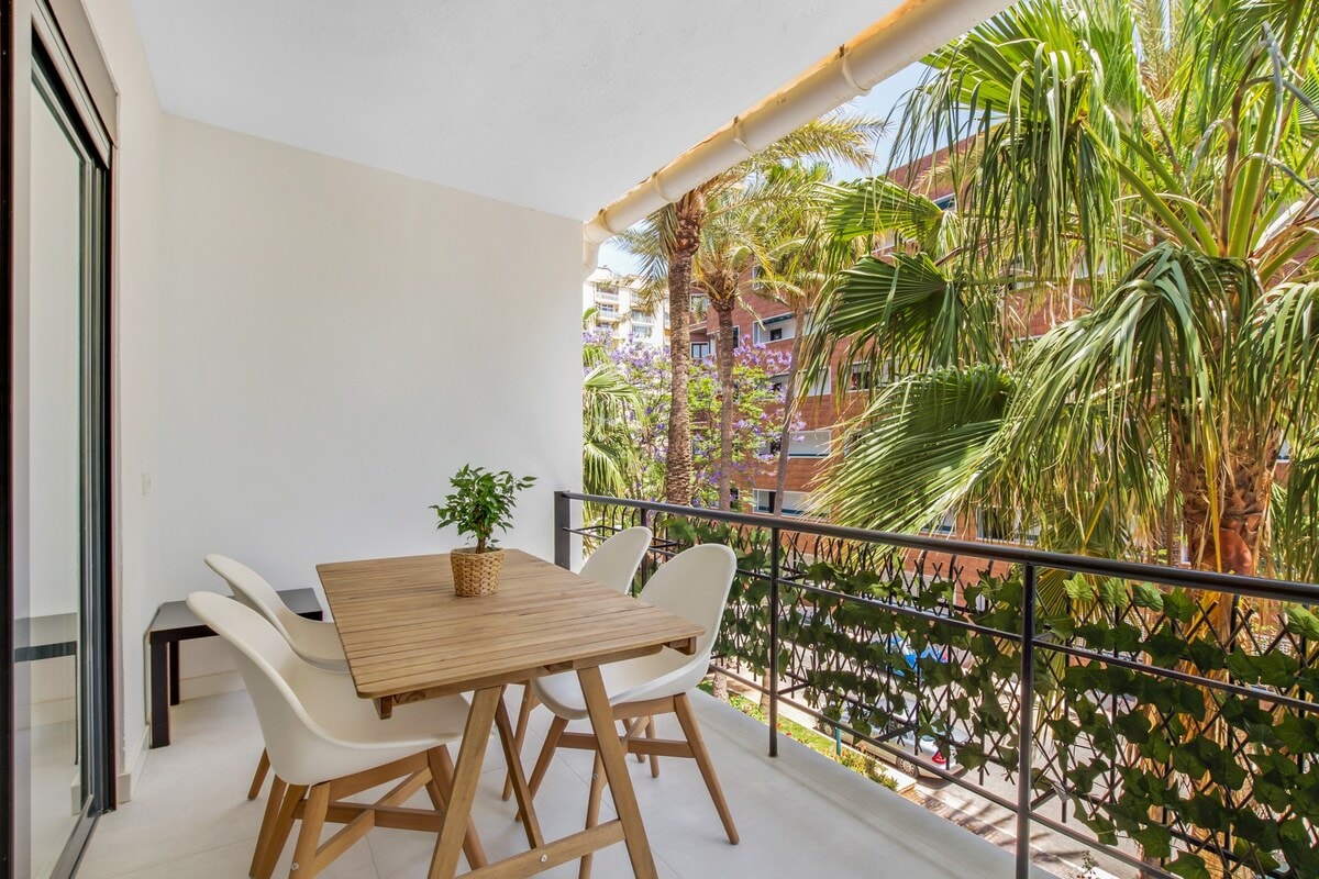 A private terrace is shown, featuring a wooden table surrounded by white chairs. Lush palm trees are visible in the background, adding a touch of greenery to the space. Natural light fills the area, highlighting the modern design and outdoor dining option.