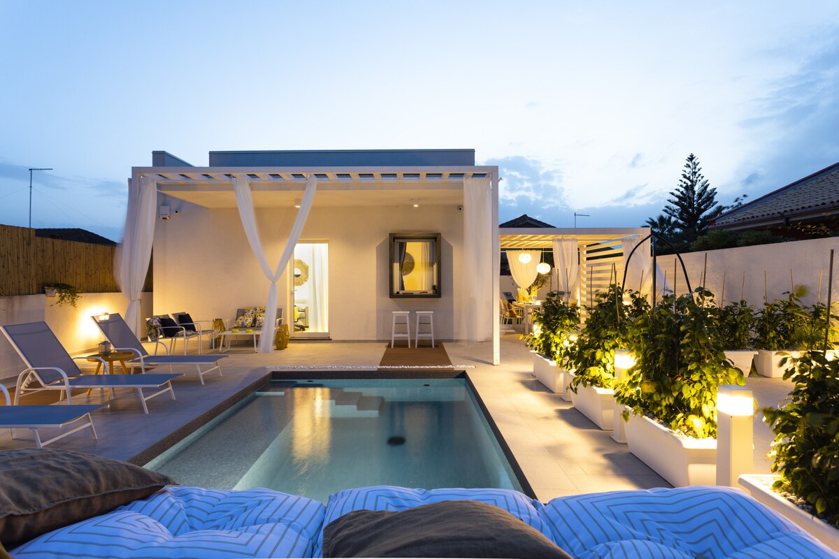 The villa exterior is visible at twilight, featuring a private swimming pool surrounded by lounge chairs. Soft lighting illuminates the outdoor area, highlighting potted greenery. A covered terrace extends from the villa, providing a shaded space that opens to the inviting interior.