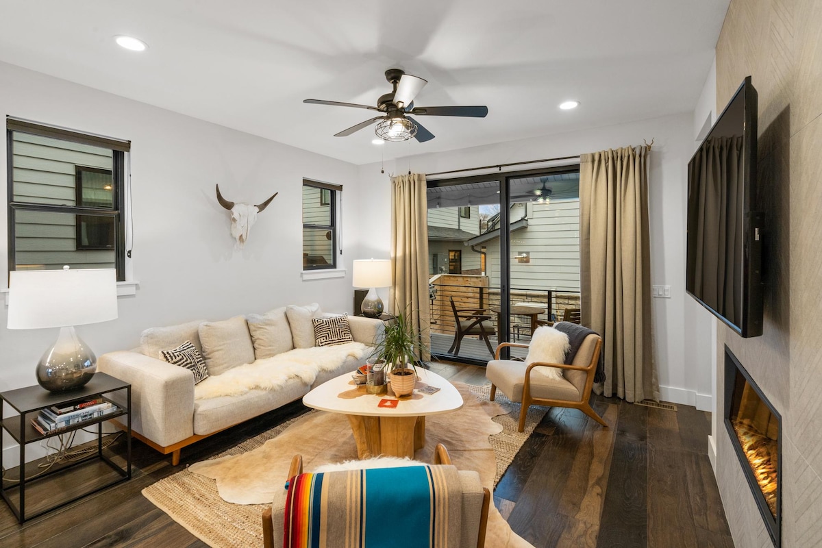 The living room is designed with a neutral palette, featuring a spacious sectional sofa and a round coffee table. Floor-to-ceiling windows offer natural light and outdoor views. A ceiling fan and a sleek fireplace enhance the comfortable atmosphere. Soft, textured rugs anchor the space.