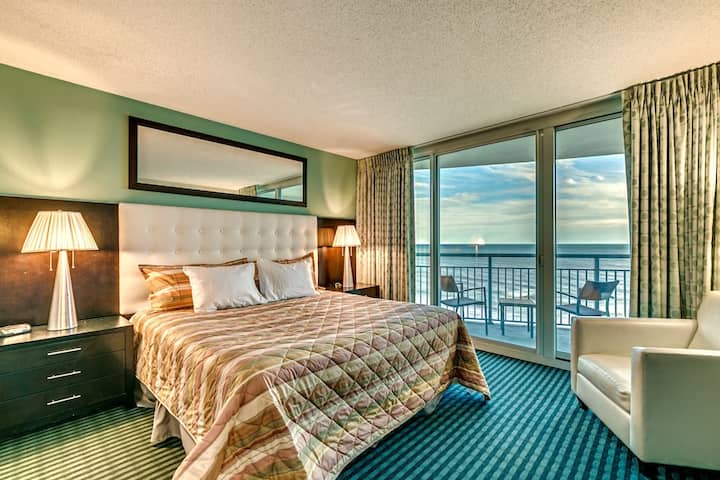 Елегантна спалня с плюшено легло с ватирана табла, топло нощно осветление и прозорци от пода до тавана, които се отварят към балкон със зашеметяваща гледка към океана, създавайки спокойна атмосфера за отдих.