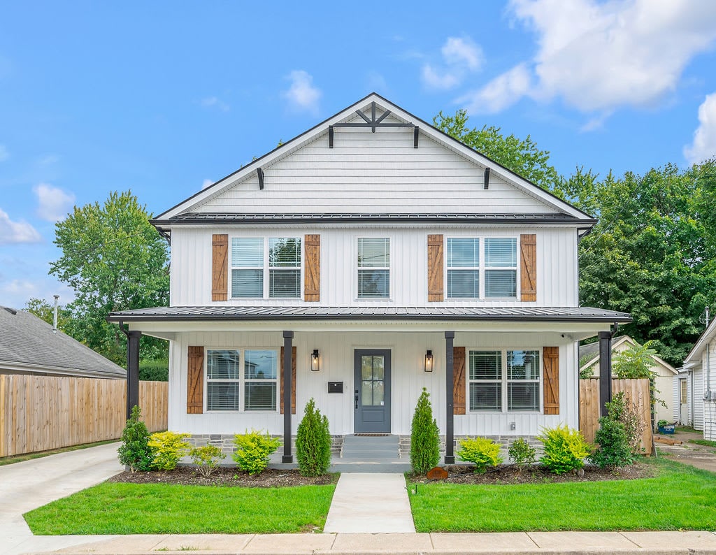 A modern farmhouse exterior is showcased, featuring a two-story structure with a symmetrical design. Wooden shutters frame the large windows, and fresh green landscaping is visible in the front yard. A wide driveway leads up to the entrance, complemented by a well-maintained lawn.