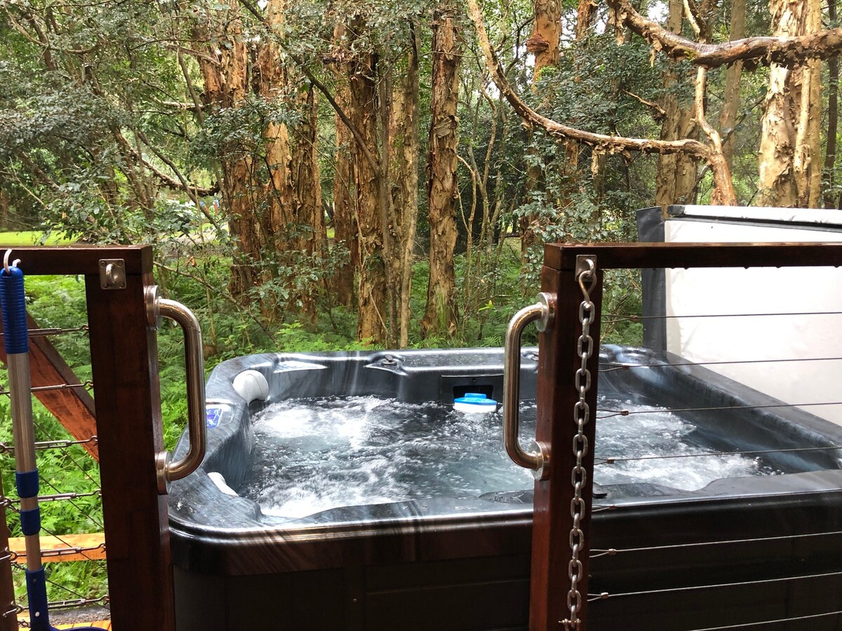 A private outdoor hot tub is surrounded by lush greenery, offering a serene view of the forest. The wooden deck includes protective railings, enhancing privacy while allowing guests to enjoy the natural surroundings.