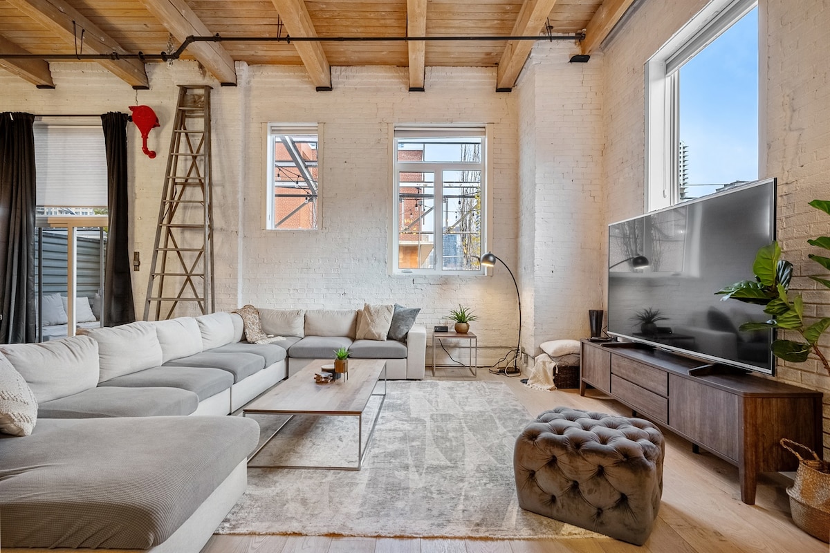 A spacious living area features a large sectional sofa arranged around a low coffee table. A flat-screen television is mounted on a light wood media console. Large windows fill the space with natural light, and a potted plant adds a touch of greenery to the neutral color palette.