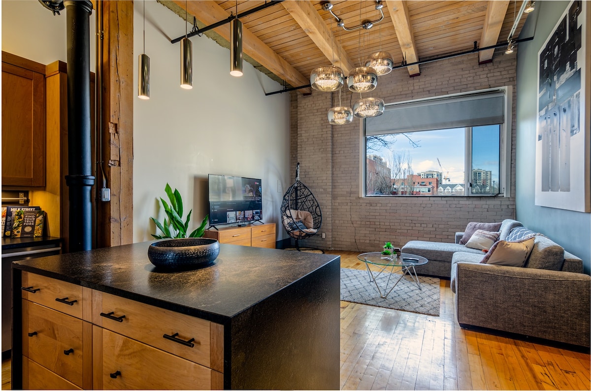 The living area features exposed wooden beams and a large window providing natural light. A comfortable gray couch faces a modern television, while a circular coffee table sits atop a textured rug. A green plant adds a pop of color next to a stylish hanging chair.