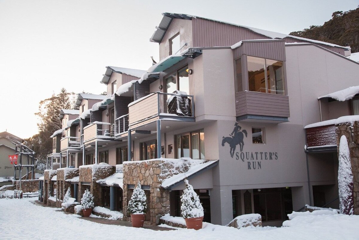A multi-story building with a modern design is showcased in a snowy landscape. Balconies line the exterior, offering access to outdoor spaces. Snow-covered potted plants enhance the welcoming entrance, while the name 'Squatter's Run' is prominently displayed on one wall.