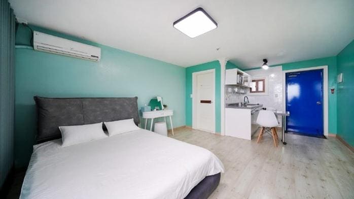 A spacious room features a large bed with neutral bedding, complemented by bright turquoise walls. A small desk and chair are provided, along with a kitchenette that includes a sink and cooking area. A bold blue door adds a striking contrast to the space.