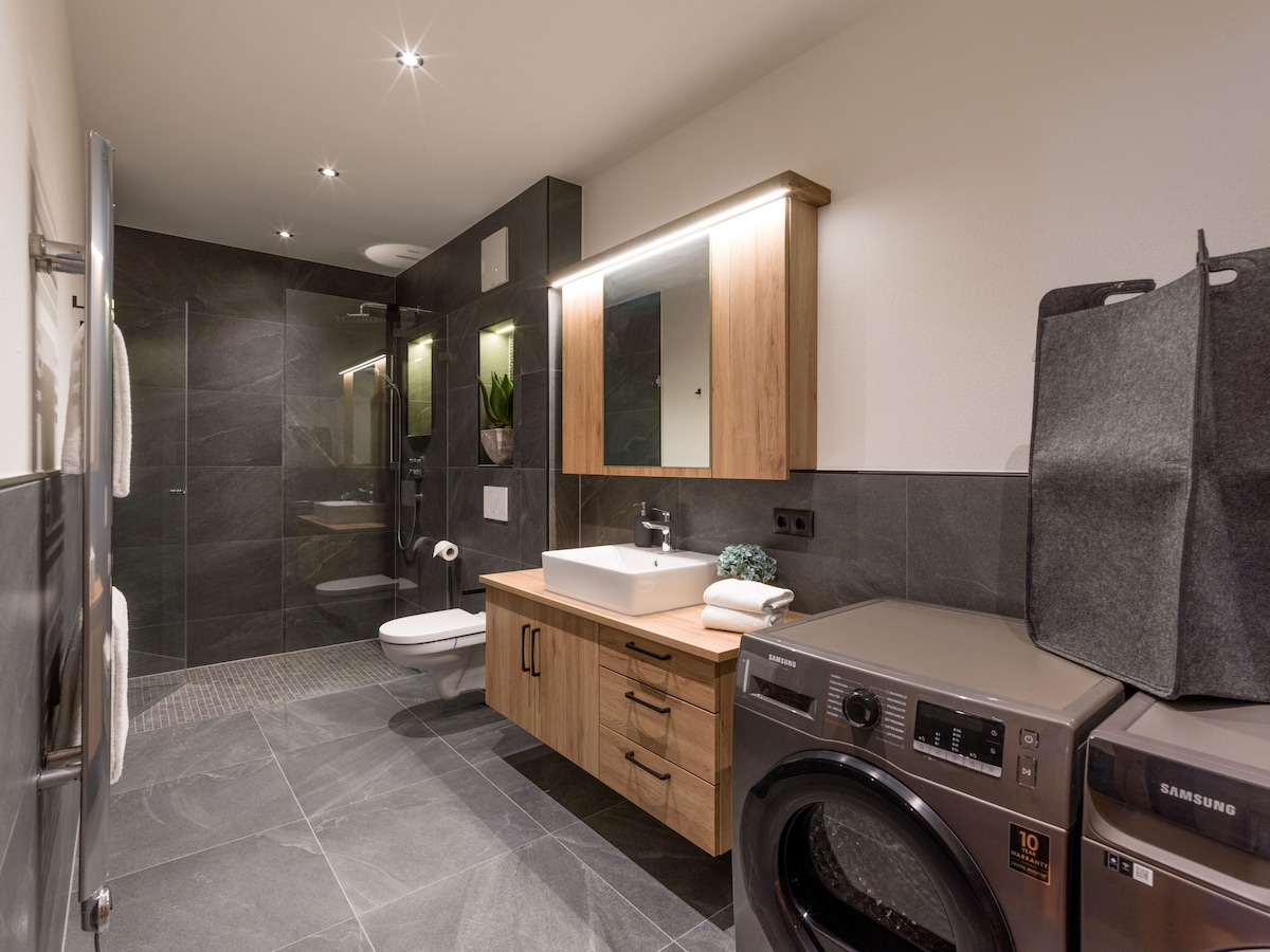 A modern bathroom is equipped with a glass shower cubicle and sleek wooden cabinetry. A wall-mounted mirror reflects ample light, while a spacious wash basin is accompanied by plush towels. A washing machine is situated beside the cabinetry, providing convenient laundry options.