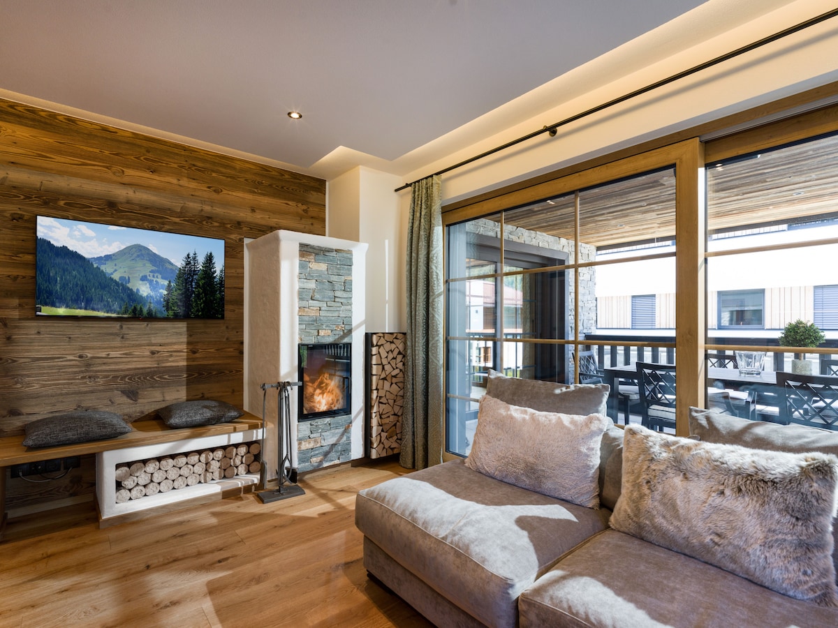 A cozy living area features a large corner sofa, complemented by textured cushions. A flat-screen TV is mounted on a wooden accent wall, while a modern fireplace sits nearby, surrounded by neatly stacked firewood. Glass doors lead to a spacious outdoor patio.
