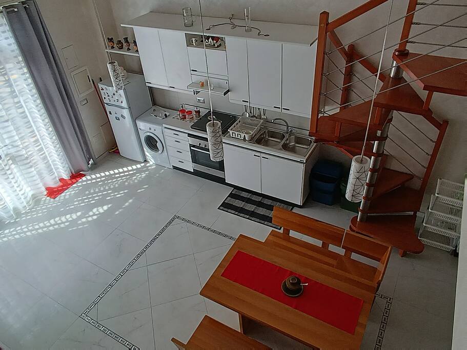 An open living space displays a modern kitchen equipped with essential appliances, including a refrigerator and a washing machine. A wooden dining table with chairs is positioned near a staircase leading to the lofted bedroom. Natural light filters through the sheer curtains, creating a bright ambiance.