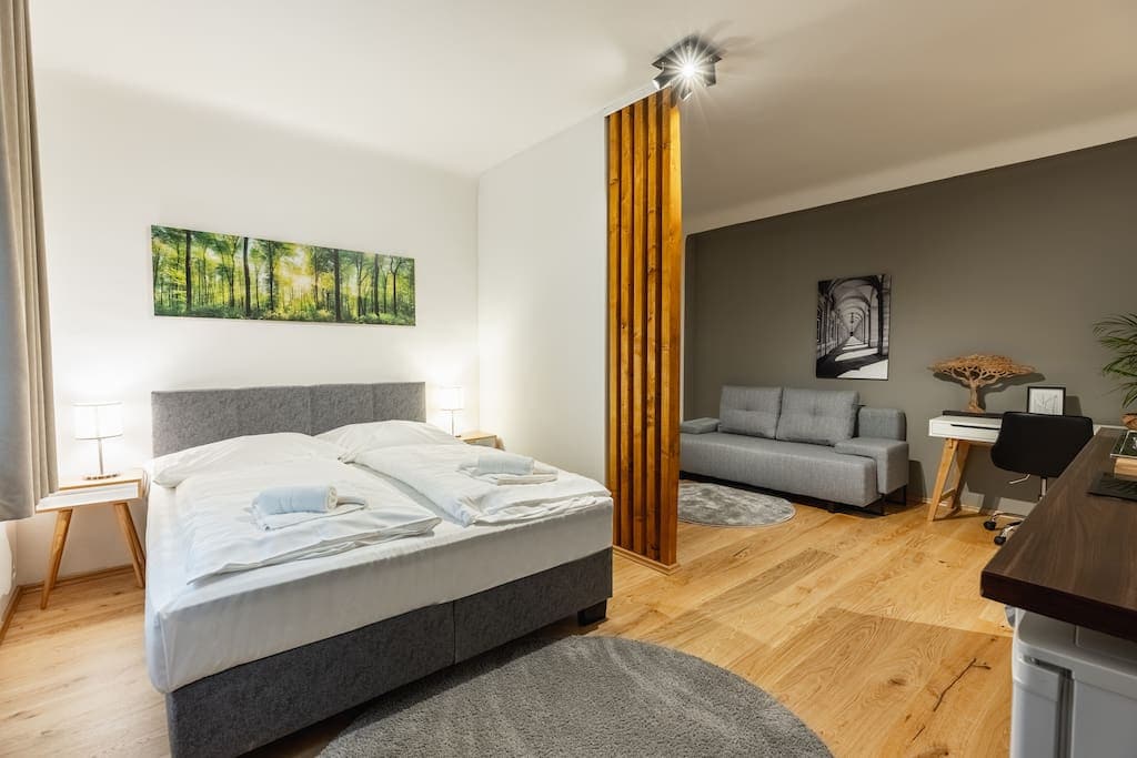 A studio apartment is presented with a queen-size bed adorned with crisp linens. Adjacent, a comfortable couch is situated beside a workspace featuring modern decor. Natural wood accents and soft lighting enhance the warm atmosphere, while art pieces are displayed on the walls.